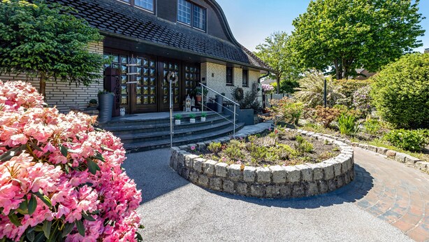 A building with a curved roof, a stone pathway, and a landscaped garden with pink flowers. - Apartment 'Storchenallee' (Burg (Dithmarschen))
