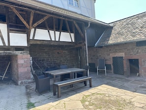 Un patio con una mesa de piedra, dos sillas, un banco y un edificio parcialmente visible con vigas de madera a la vista y paredes de piedra.