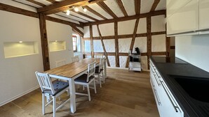 Un coin repas avec une table et des chaises en bois, une cuisine équipée d’une cuisinière et un plafond voûté avec des poutres apparentes.