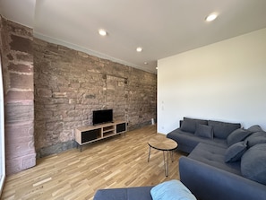 Una sala de estar con una pared de piedra, un televisor de pantalla plana sobre un soporte de madera, un sofá gris y una mesita redonda pequeña.
