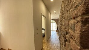 Un couloir doté d’un plancher en bois, d’un mur en pierre et d’une porte donnant sur une pièce contenant un lit et une fenêtre.
