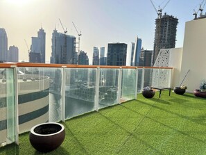 A rooftop terrace with artificial grass, two large planters, a glass railing, and a city skyline in the background. - 1BR Penthouse in Millenium Binghatti  (Dubai)