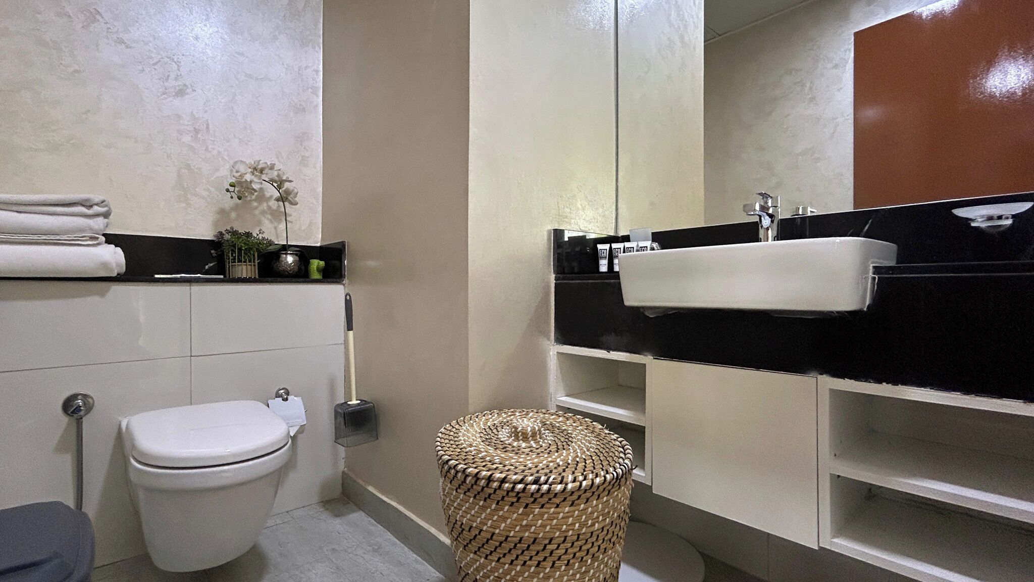 Une salle de bain avec des toilettes blanches, un lavabo noir et une poubelle en osier.