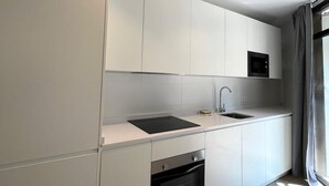 Une cuisine moderne avec des meubles blancs, une plaque de cuisson à induction noire, un four encastré et une fenêtre avec des rideaux.
