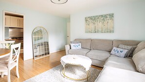 A modern living room with a sofa, ottoman, and dining table. There is a kitchen area with cabinets and a stove. - Quiet and Cosy 2-bed in Wimbledon (London)