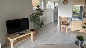 Ein Wohnzimmer mit Fernseher, einem Holztisch, einer Topfpflanze und einer Küchenzeile mit Esstisch und Stühlen.
