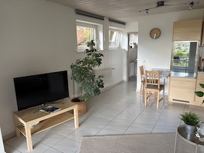 A living room with a TV, a wooden table, a potted plant, and a kitchen area with a dining table and chairs.