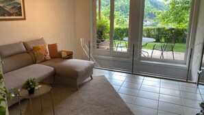 Ein modernes Wohnzimmer mit einem Ecksofa, einem kleinen Beistelltisch mit Pflanze und einer großen Schiebeglasdoor, die zu einer Außenpatio führt.