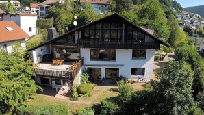 Ein zweistöckiges Haus mit Balkon, Terrasse und Blick auf einen bewaldeten Hügel.