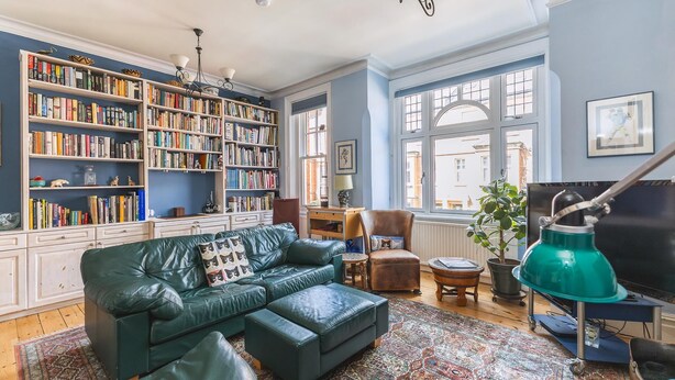 A cozy living room with a green leather sofa, a bookshelf filled with books, a television, and a large window with a view of buildings outside. - Stylish Hampstead Retreat by the Heath (London)