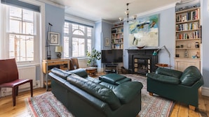 A living room with a sofa, armchair, fireplace, bookshelf, and artwork. - Stylish Hampstead Retreat by the Heath (London)