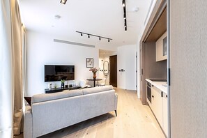 A modern living room with a sofa, a flat-screen TV, a small table with a vase, and a kitchen area with cabinets and a dishwasher. - Elegant Flat in Notting Hill's Heartbeat (London)
