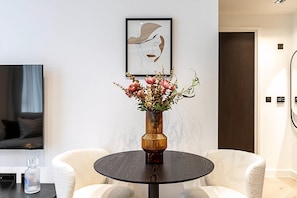 Una sala de estar moderna con una mesa redonda, dos sillas blancas, un televisor y una obra de arte enmarcada en la pared.