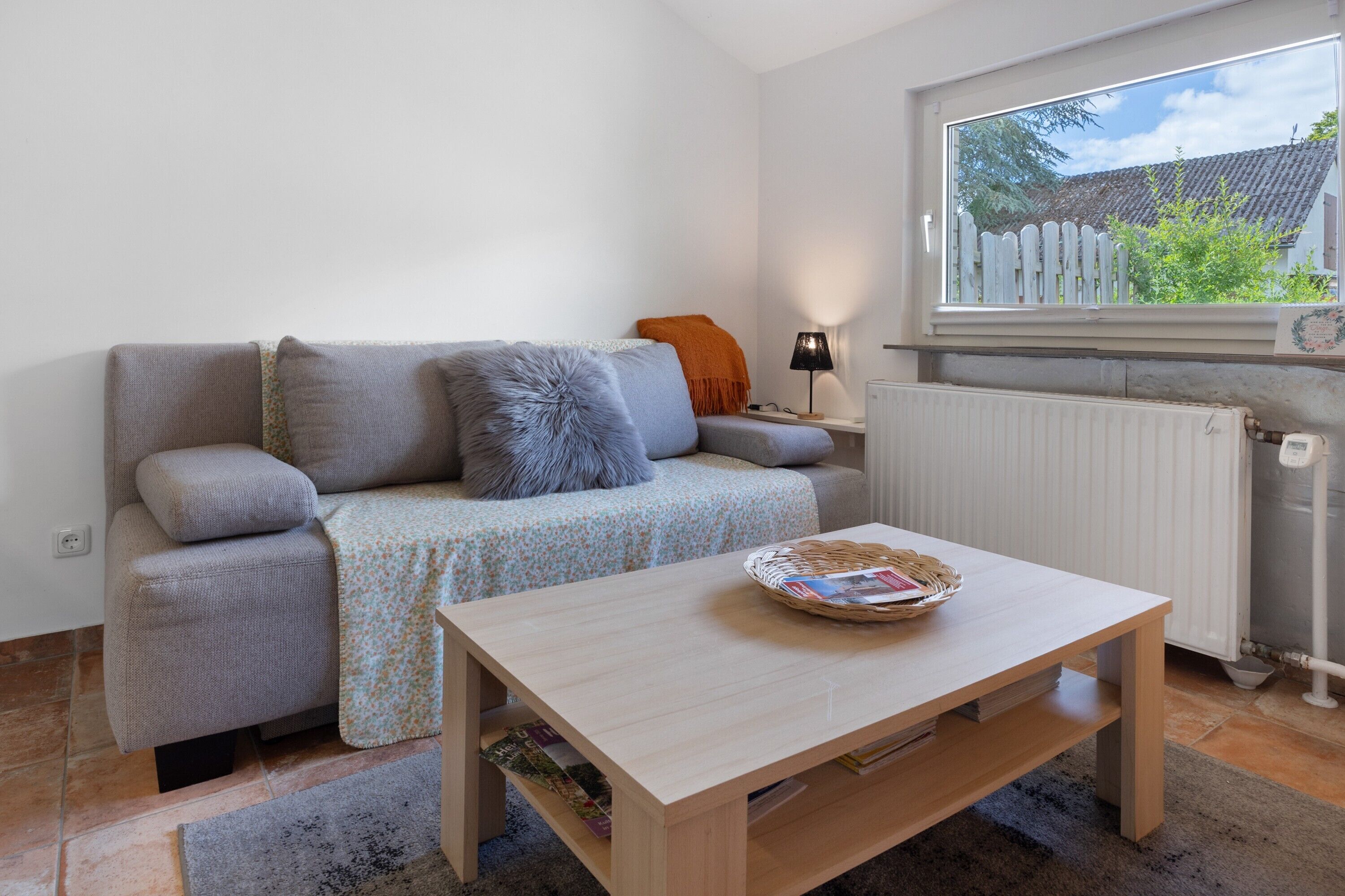 A living room with a sofa, a table, a lamp, and a window with a view of a house and trees.