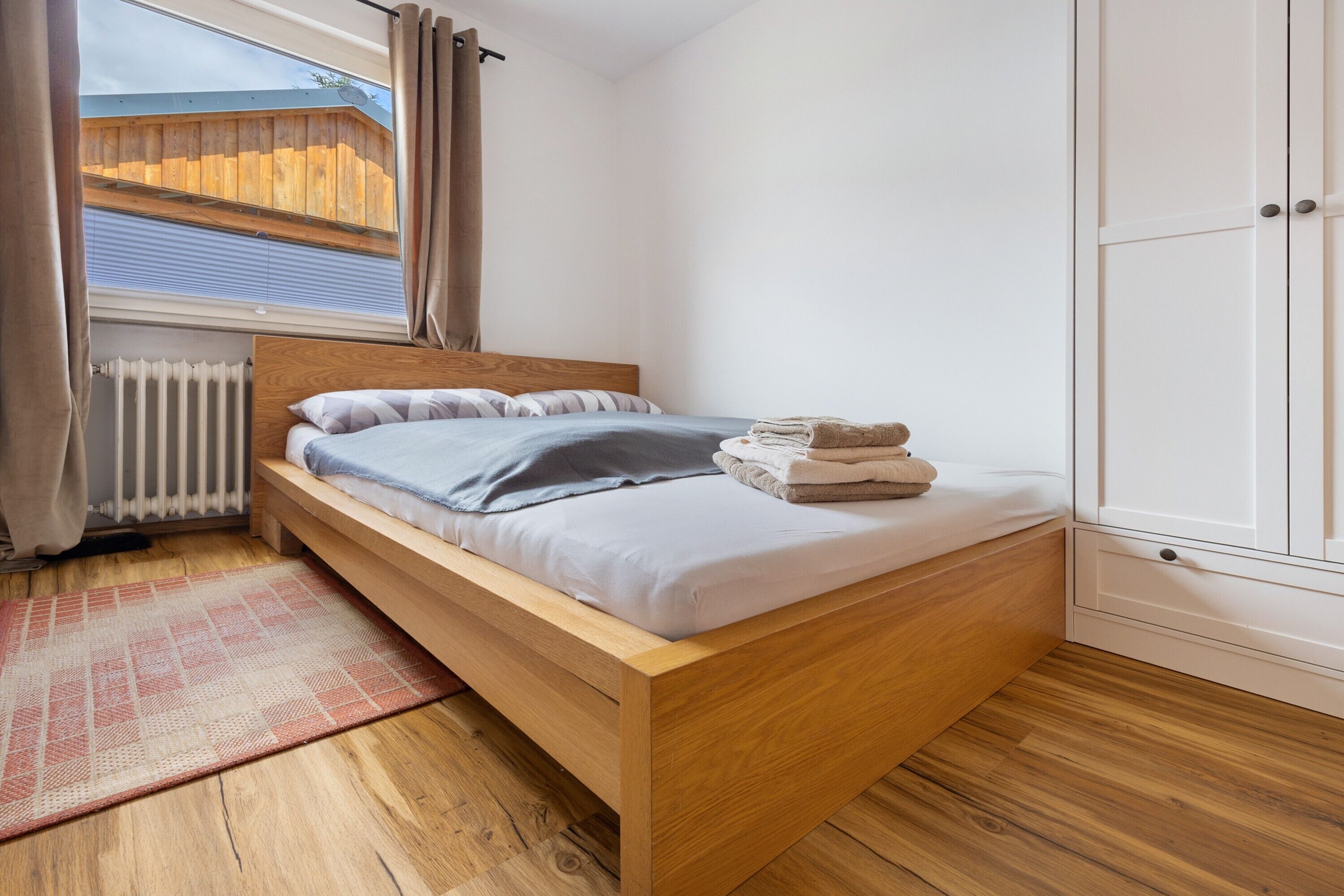 Ein Einzelbett mit Holzrahmen, ein Fenster mit Vorhängen, ein Heizkörper und ein Kleiderschrank.