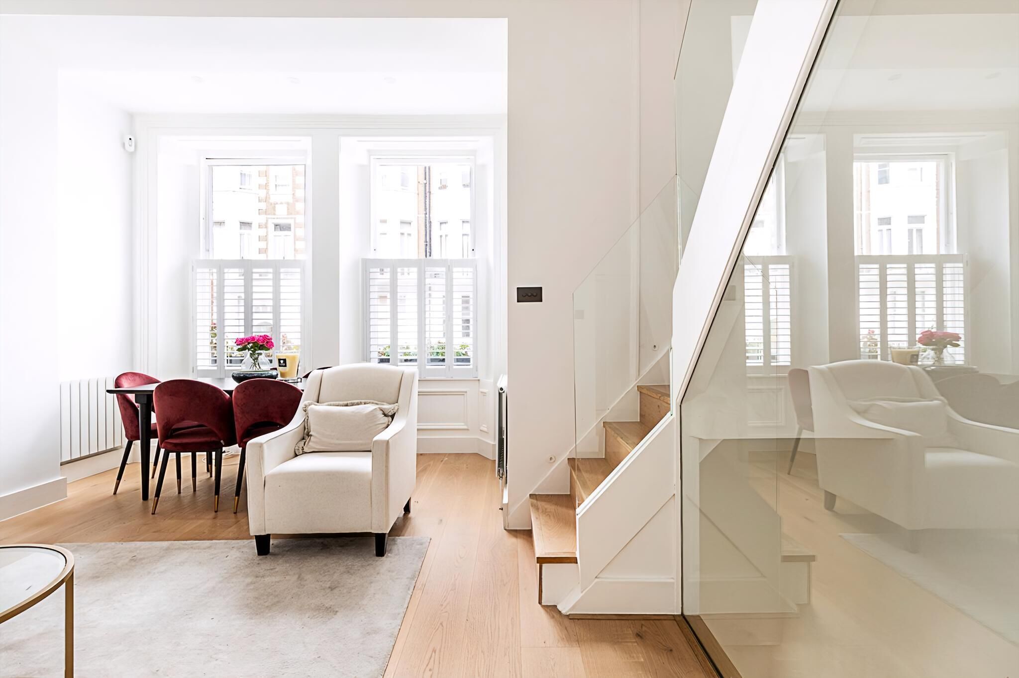 A modern living room with a white sofa, red chairs, a dining table with chairs, and a staircase with glass railings.