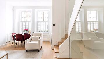 A modern living room with a white sofa, red chairs, a dining table with chairs, and a staircase with glass railings.