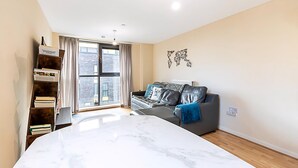 A modern living room with a large bed, a grey sofa, a bookshelf, and a window with curtains.