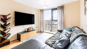 A modern living room with a large flat-screen TV, a bookshelf, a sofa, and a view of the city through a large window.