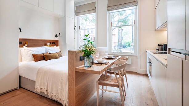 A modern, compact bedroom with a wooden headboard, a small dining area with a table and chairs, and a kitchenette with a refrigerator and stove. - The Philbeach Three (London)