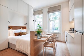 Een moderne, compacte slaapkamer met een houten hoofdeinde, een klein zitehoekje met een tafel en stoelen, en een kitchenette met een koelkast en fornuis.