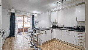 Una cocina moderna con armarios blancos, encimera negra y electrodomésticos de acero inoxidable. Cuenta con un comedor con mesa y sillas, y vistas al exterior a través de un ventanal.