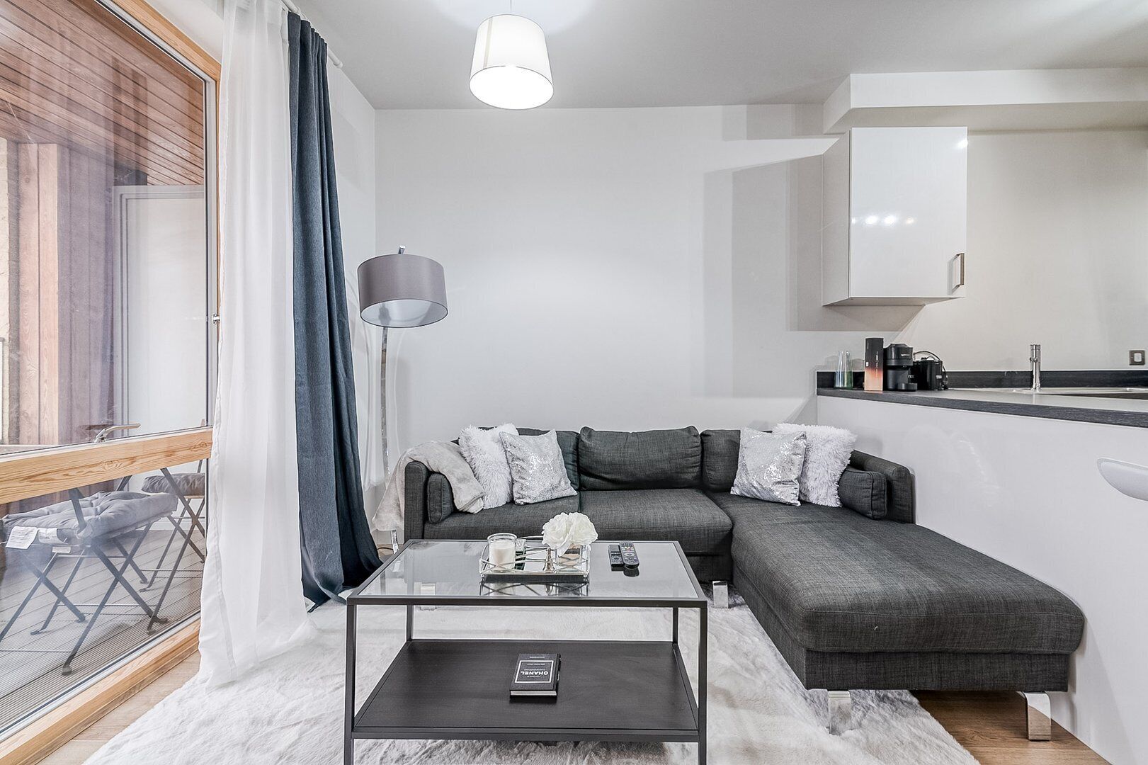 A modern living room with a sectional sofa, a glass coffee table, and a kitchen area with a white countertop and cabinets.