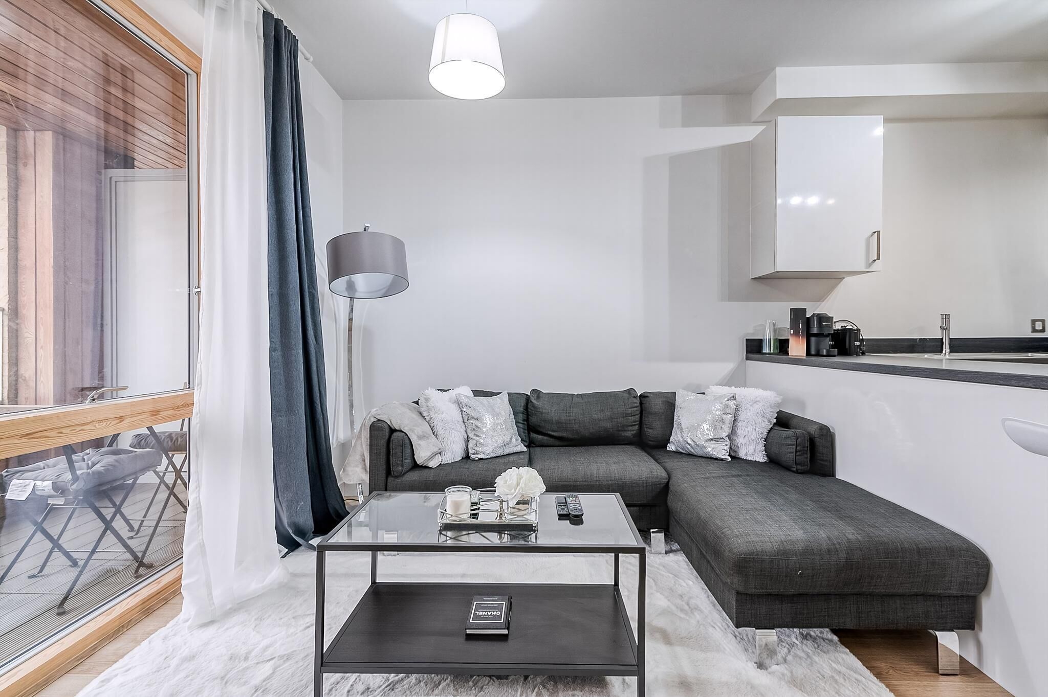 A modern living room with a glass coffee table, a grey sofa, and a large window with curtains.