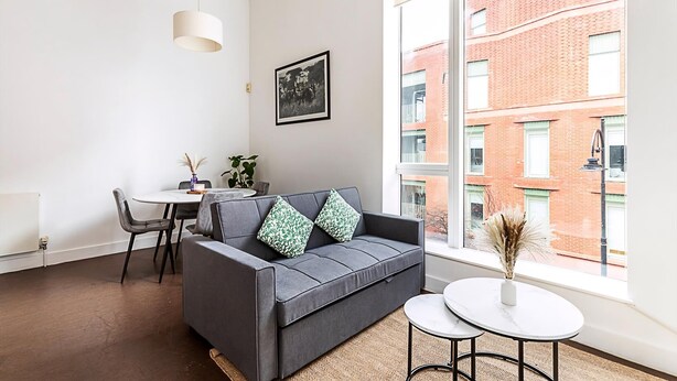 Ett modernt vardagsrum med en grå soffa, ett litet matbord och ett stort fönster med utsikt över en byggnad med tegelstensfasad.