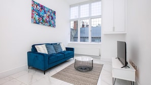 Una sala de estar moderna con un sofá azul, una mesa de centro redonda, un televisor de pantalla plana sobre un soporte blanco, un cuadro abstracto grande y una ventana con vistas a edificios.