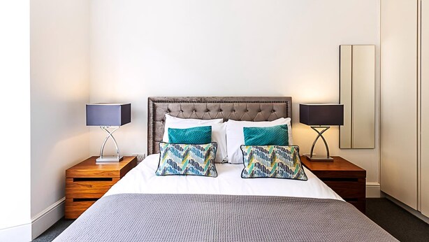A neatly arranged hotel room with a tufted headboard, a large bed with patterned pillows, two bedside tables with lamps, and a large mirror on the wall. - Stylish Two-bed Flat Near Oxford Circus (London)