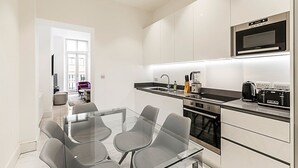 A modern kitchen with a glass dining table, grey chairs, and stainless steel appliances.