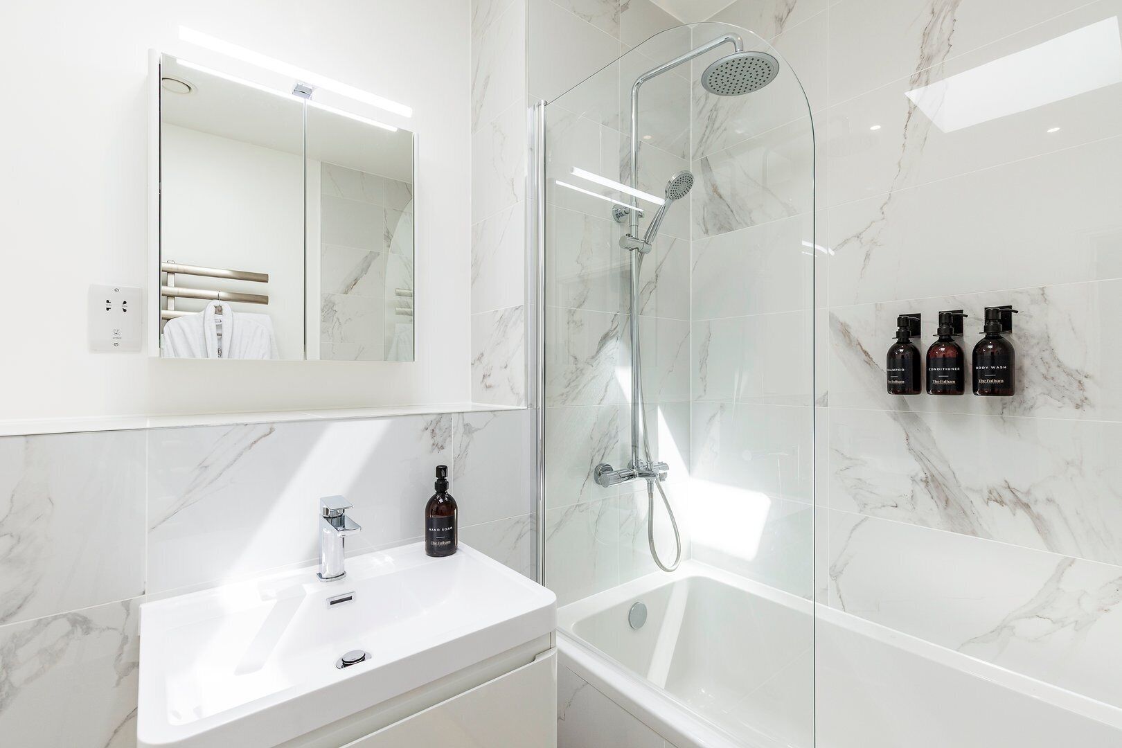 A modern bathroom with a glass-enclosed shower, a white sink, and a mirror above it. There are three bottles of hand soap and body wash on the wall.