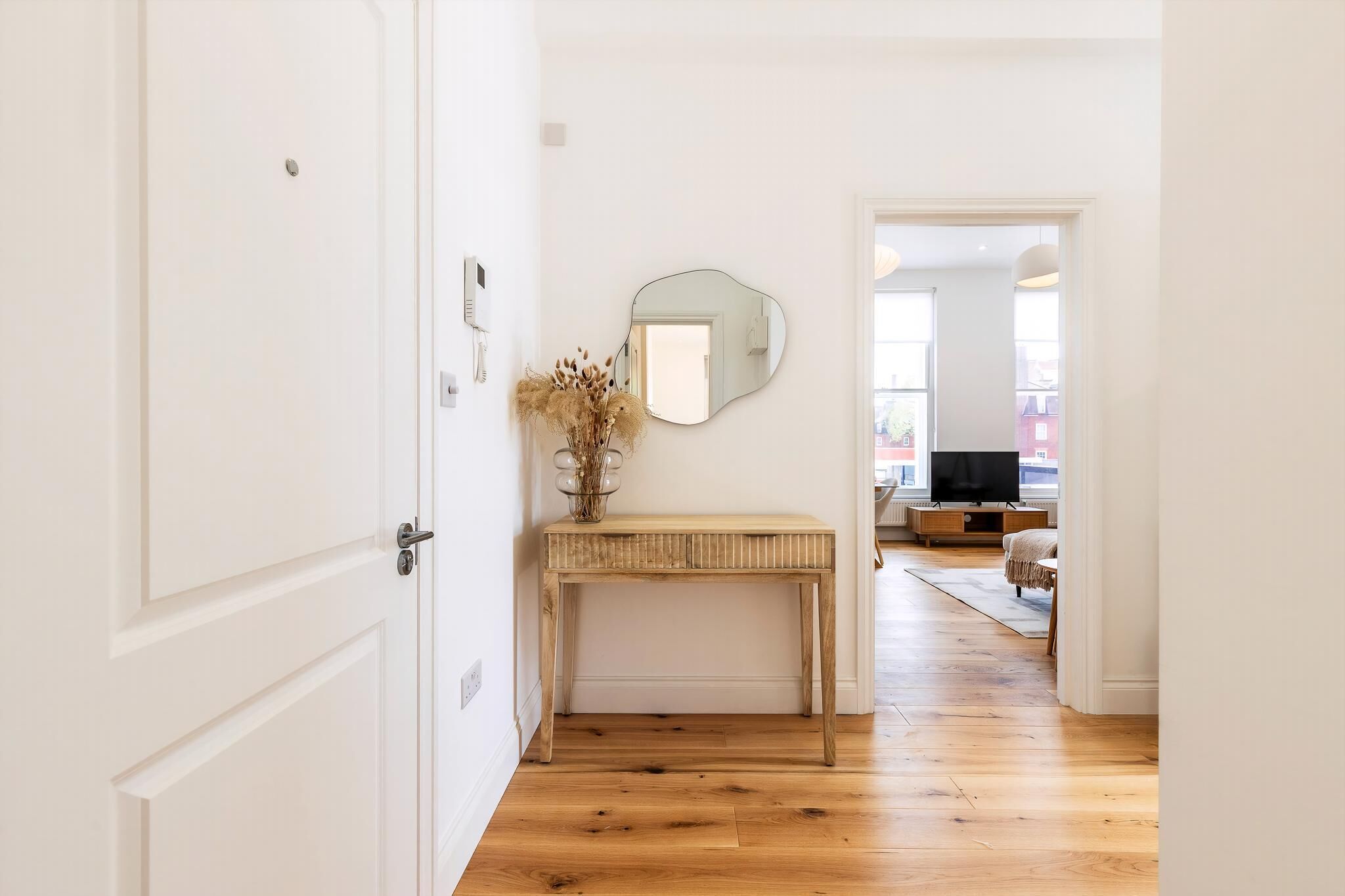 Un couloir avec un plancher en bois, une porte blanche, une console surmontée d’un miroir et un vase rempli de fleurs séchées.