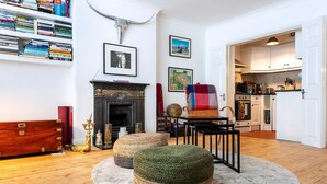 Una sala de estar con chimenea, otomanes, una estantería y una cocina visible a través de una puerta abierta.