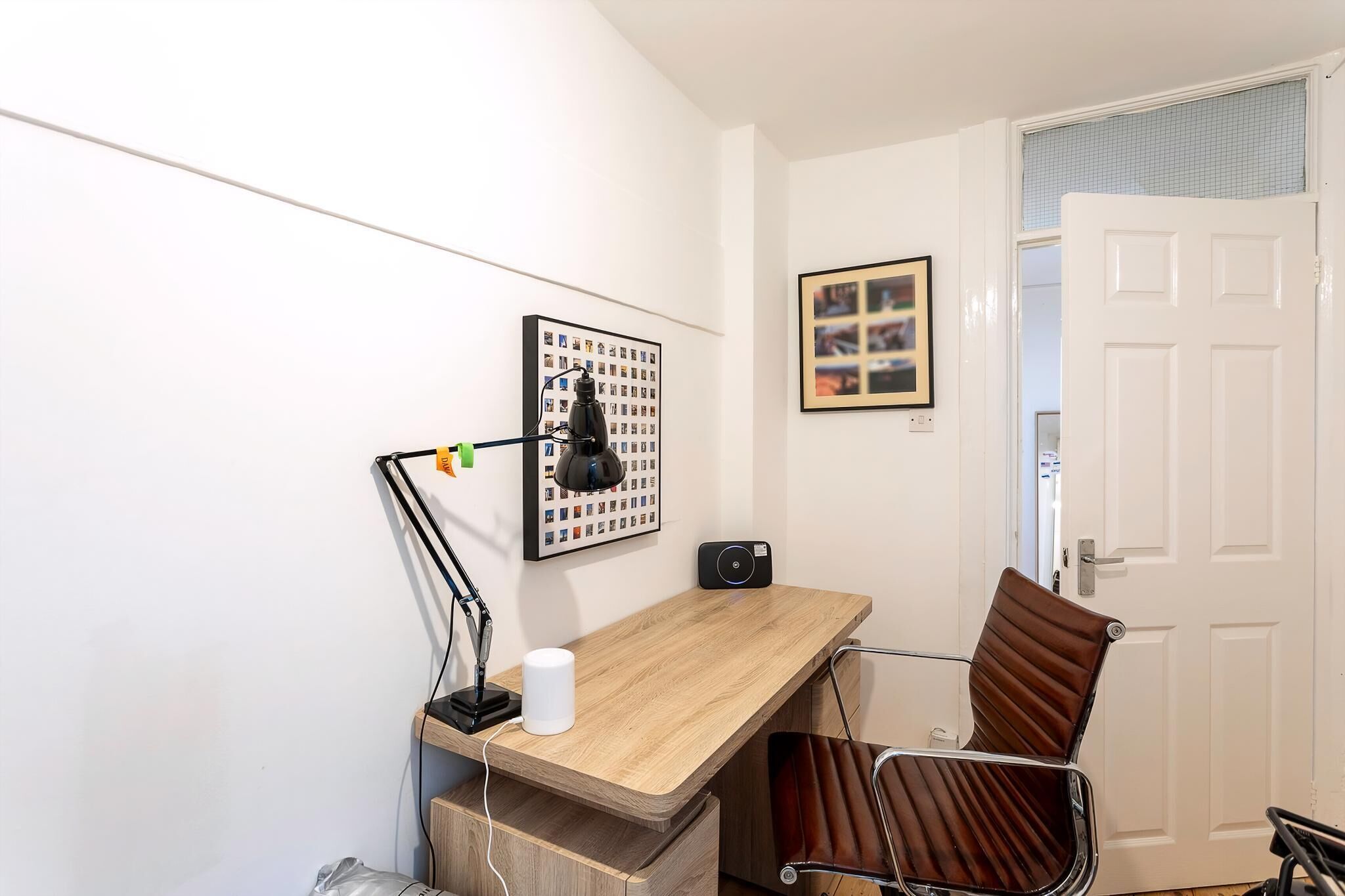 A small office space with a wooden desk, a brown leather chair, a black desk lamp, a framed photo collage, and a white door.