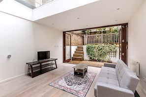 A modern living room with a sofa, TV, and coffee table, a patterned rug, and a view of a brick staircase and garden. - Beautiful Modern 4BR House in Central London (London)