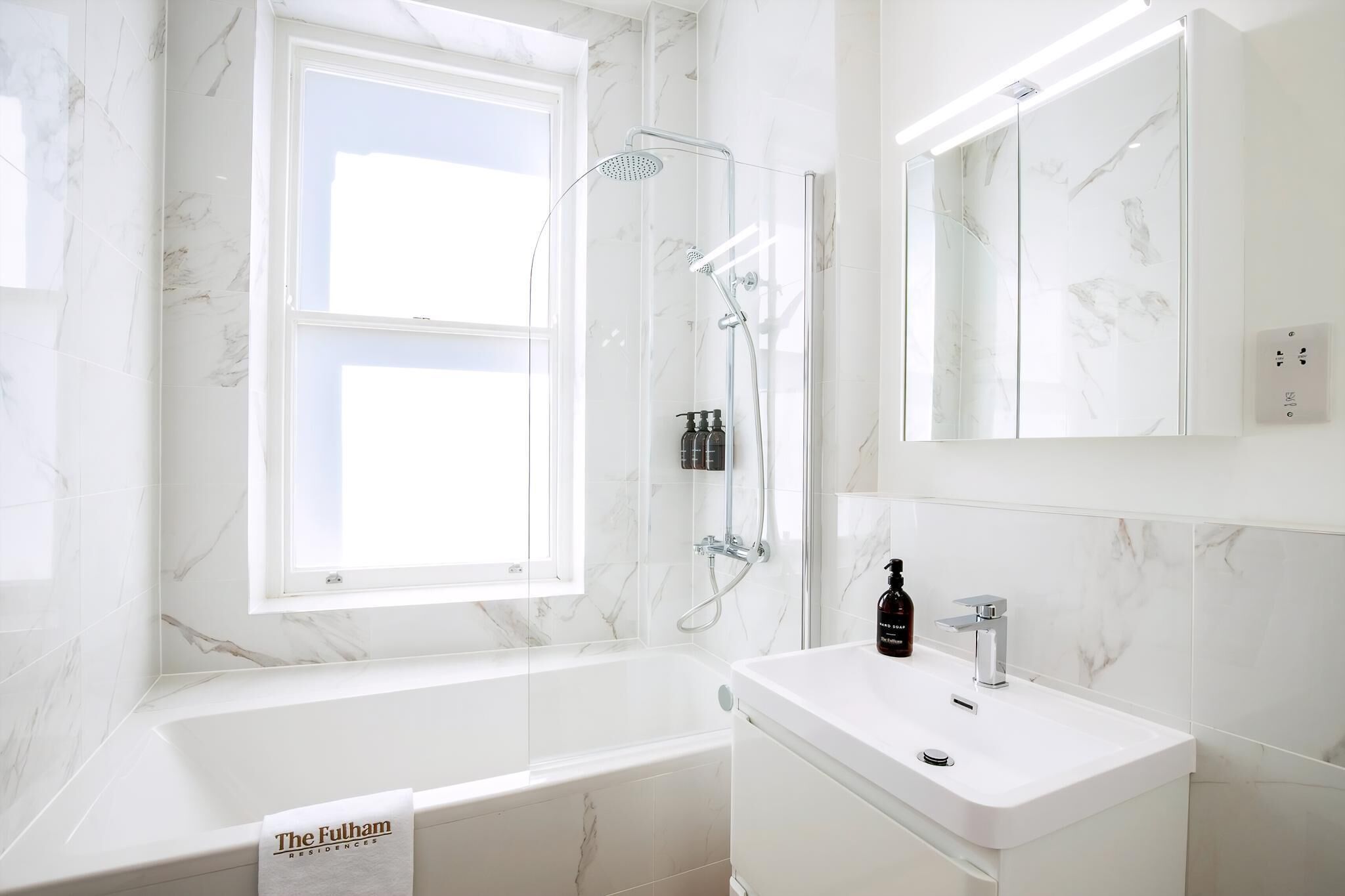 A modern bathroom with a white marble wall, a large window, a bathtub, a sink, and a glass-enclosed shower.