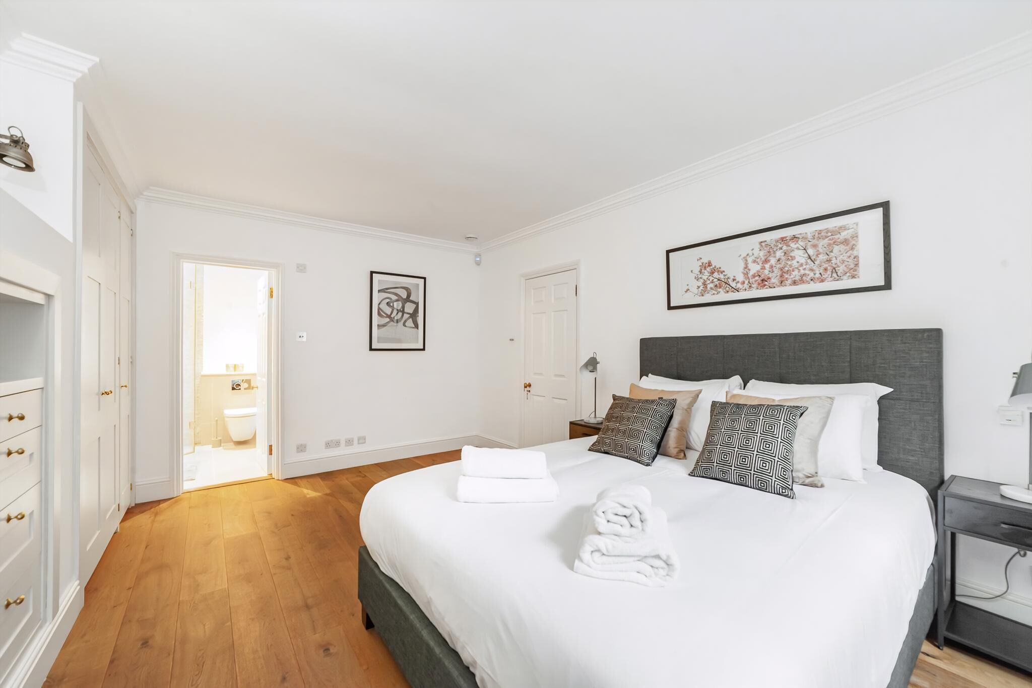 Une chambre avec un grand lit, du linge de lit blanc et une tête de lit grise. On y trouve deux tables de chevet avec des lampes et un tableau encadré au mur.