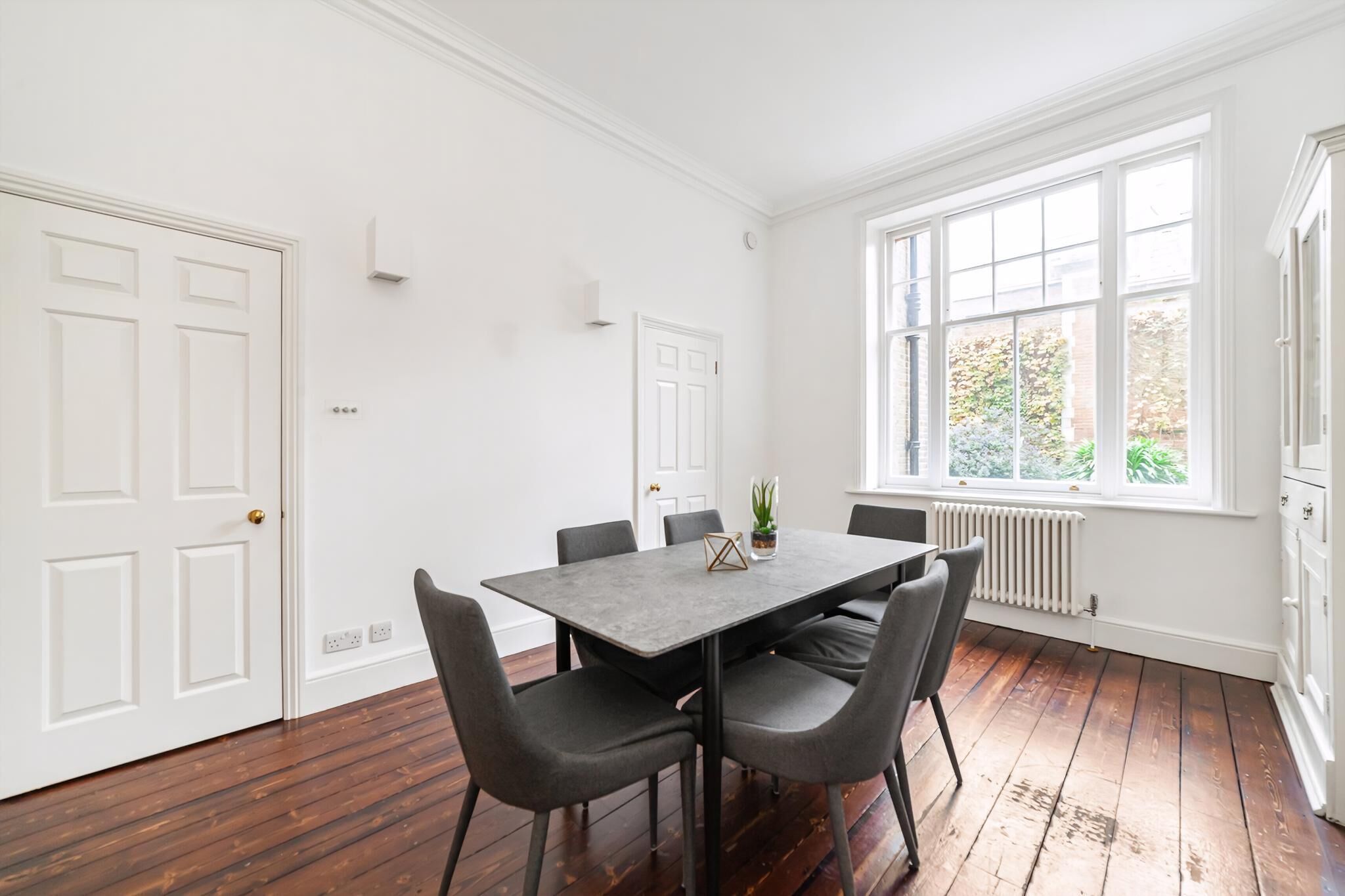 A dining area with a rectangular table, six chairs, a potted plant, and a large window with a view of greenery.
