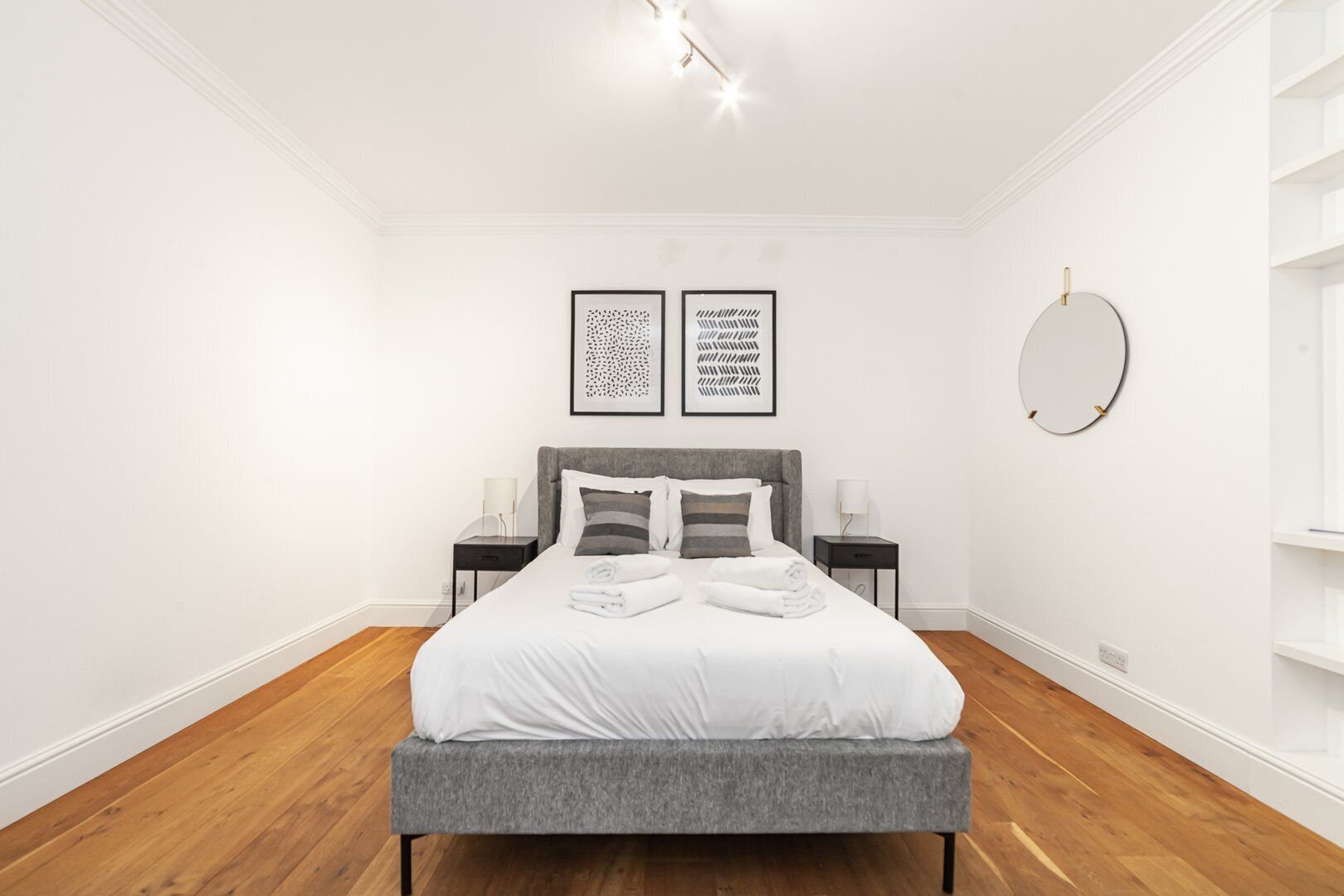 Ein modernes Schlafzimmer mit einem großen Bett, zwei Nachttischen, einem runden Spiegel und zwei gerahmten Kunstwerken an der Wand.