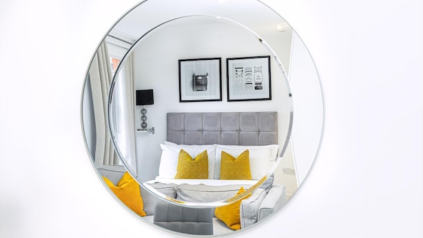 Un espejo circular que refleja un dormitorio con una cama, almohadas y dos cuadros en la pared.