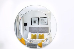 A circular mirror reflecting a bedroom with a bed, pillows, and two framed pictures on the wall. - Stylish Studio Retreat in Trendy Marylebone (London)