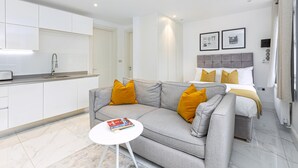 Una habitación de hotel moderna con un sofá gris, una mesita redonda pequeña, una cama con cabecera gris y una zona de cocina con armarios blancos y un lavamanos.
