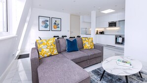 A modern living room with a grey sectional sofa, a round coffee table, and a view of the kitchen in the background.