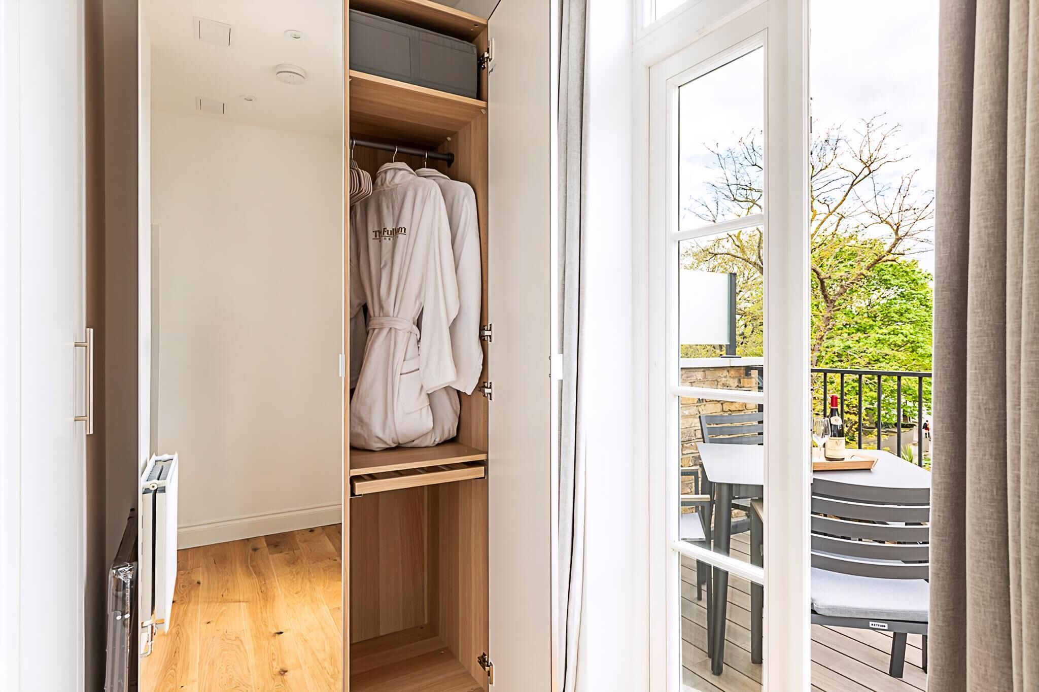 Un placard ouvert où sont suspendues des vêtements, un plancher en bois et une vue sur un balcon extérieur avec une table et des chaises.