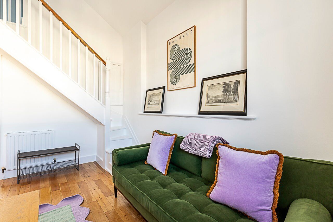 Un canapé vert avec des coussins violets, un escalier blanc et des tableaux encadrés au mur.