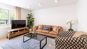 A modern living room with a flat-screen TV, a brown sofa with patterned cushions, a glass coffee table, and a large window with curtains. - 3 Bed In The Heart of Covent Garden (London)