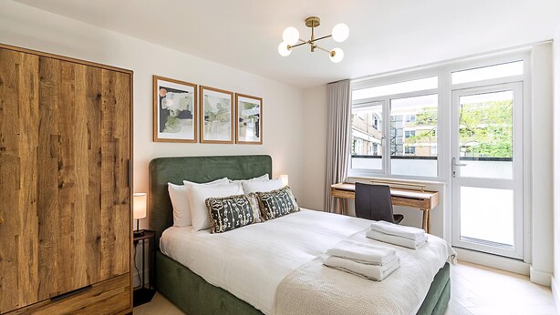 A bedroom with a green upholstered bed, white bedding, a wooden wardrobe, a desk with a chair, and a large window with a view of buildings and greenery. - 3 Bed In The Heart of Covent Garden (London)