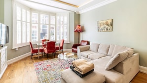 Un salon avec un canapé beige, des fauteuils rouges, une petite table et une fenêtre avec des stores.